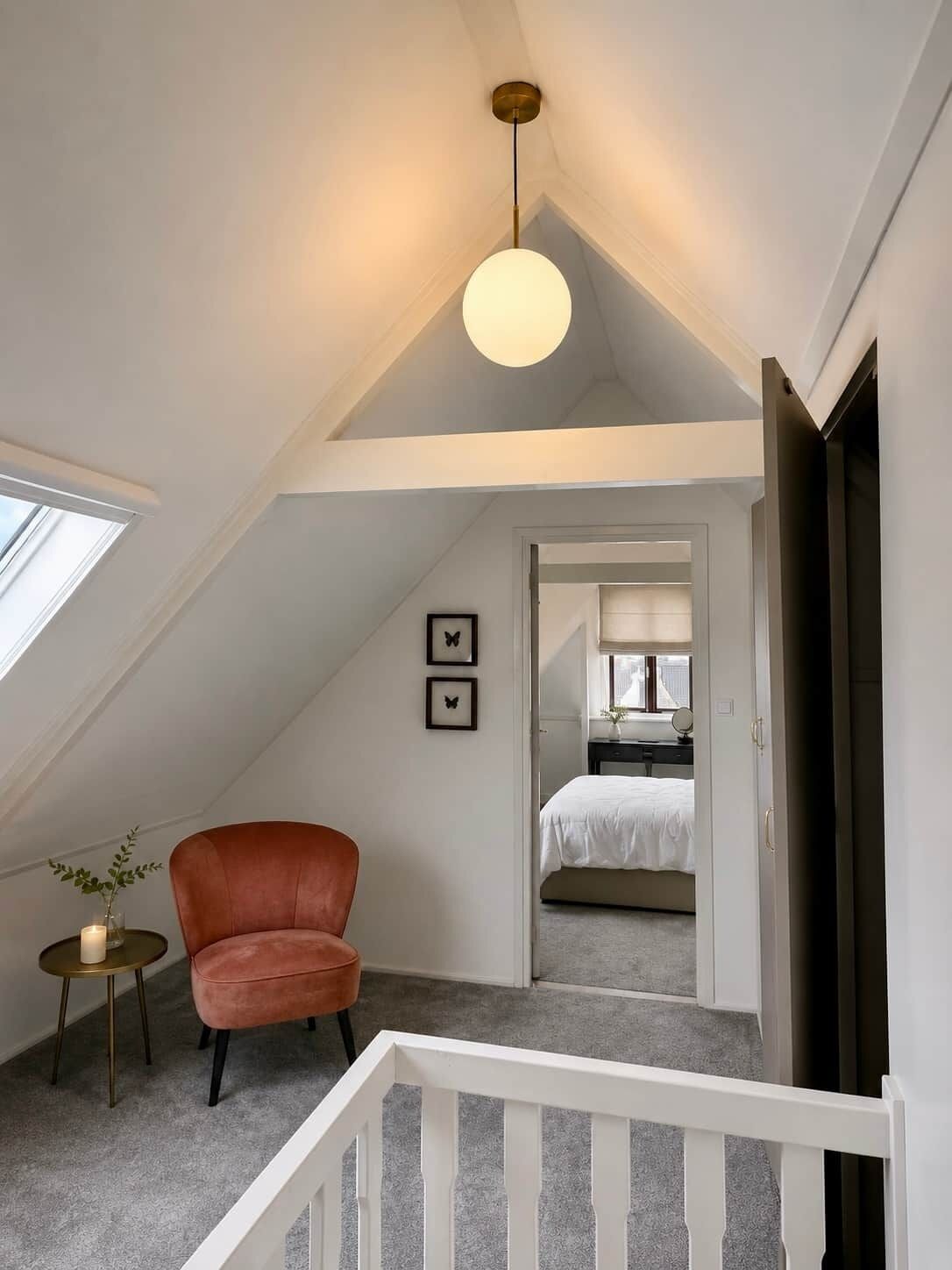Ein Schlafzimmer im Loft-Stil mit schrägem Dach, einem roten Sessel, einem kleinen Tisch mit Kerze und Blick in den angrenzenden Raum.