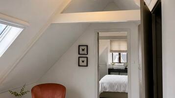 Ein Schlafzimmer im Loft-Stil mit schrägem Dach, einem roten Sessel, einem kleinen Tisch mit Kerze und Blick in den angrenzenden Raum.