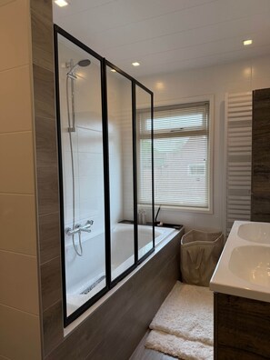 A modern bathroom with a glass-enclosed shower, a bathtub, and a window with blinds.