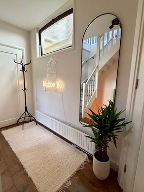 Ein Zimmer mit einem weißen Heizkörper, einem Kleiderständer, einem weißen Teppich, einer Topfpflanze und einem großen Spiegel, der eine Treppe reflektiert.