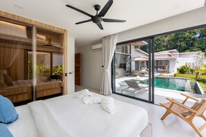 Ein modernes Schlafzimmer mit einem großen Bett, einem Deckenventilator und Blick auf einen Swimmingpool und eine Sitzgelegenheit im Freien.