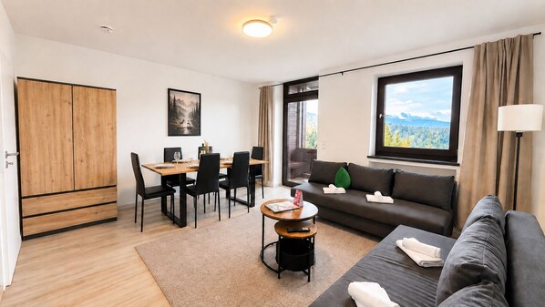 Ett modernt vardagsrum med en soffa, ett matbord med stolar, ett litet runt bord med en fjärrkontroll, ett stort fönster med utsikt över bergen och en garderob.
