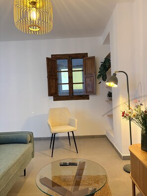 Une pièce comprenant une table ronde en verre, une chaise, un canapé, un meuble en bois, une lampe, une fenêtre avec des volets et un luminaire suspendu.