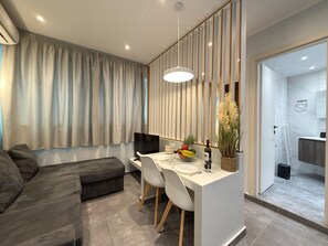 A modern living room with a sofa, a dining table with chairs, a TV, and a kitchen area visible through an open door. - SmartLiving Central Apt 3 (Thessaloniki)