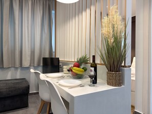 A modern kitchen with a dining area, featuring a white countertop, a bowl of fruit, a wine bottle, and a potted plant. - SmartLiving Central Apt 3 (Thessaloniki)