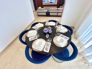 Comedor con una mesa redonda dispuesta para seis personas, rodeada de sillas azules, y un sofá y televisor al fondo.