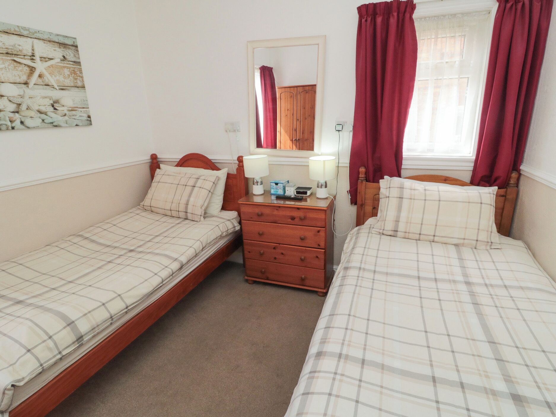 A room with two single beds, a wooden dresser, a mirror, and a window with red curtains.