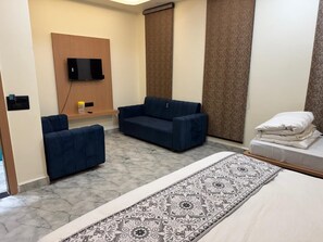 Habitación de hotel con cama, sofá azul, sofá más pequeño, alfombra estampada, televisor montado en la pared y ventana con cortinas.