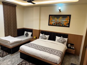A hotel room with two beds, a painting on the wall, a ceiling fan, and a nightstand with a kettle. - Hotel Shri Radhavan (Mathura)