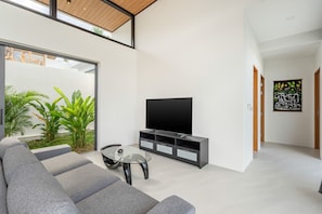 Ein modernes Wohnzimmer mit Sofa, Fernseher, Unterhaltungseinheit, einem Glastisch und Blick ins Grüne.