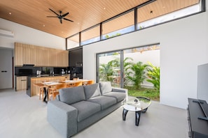 Ein modernes Interieur mit einem grauen Sofa, einem Esstisch, Stühlen, einer Küchenzeile und großen Fenstern mit Blick ins Grüne.