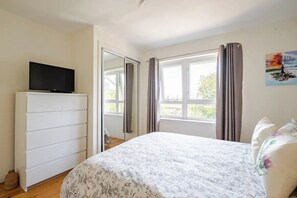 A bedroom with a bed, a TV on a dresser, a window with curtains, and a painting on the wall. - Vrbo Property (Edinburgh)