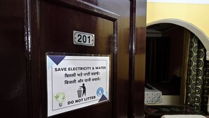 Puerta con un cartel que promueve el ahorro de electricidad y agua, un número de habitación y una cama con cubrecama blanca.