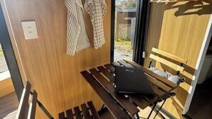 Eine Holzbank mit einem Laptop, eine Holzwand mit einem Fenster und ein Wäscheständer mit Kleidung.