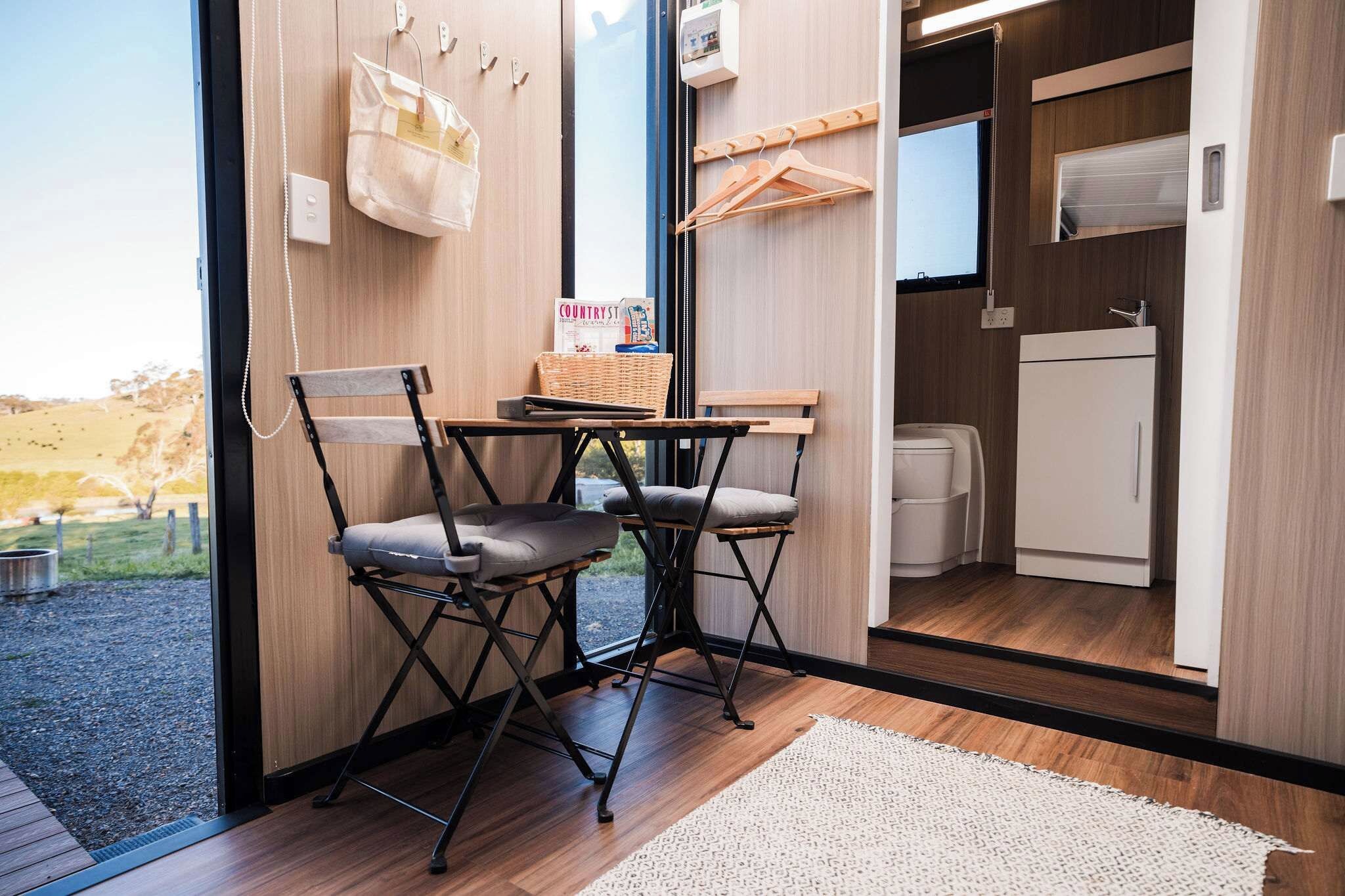 A room with a wooden floor, a table with chairs, a window with a view of a landscape, and a white refrigerator.