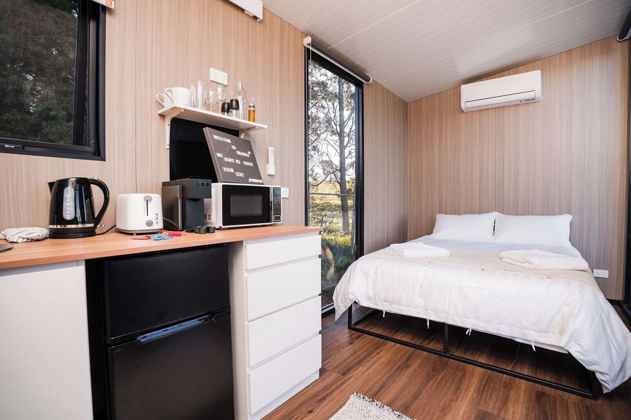 A modern bedroom with a bed, a desk with a kettle, a microwave, and a small refrigerator.