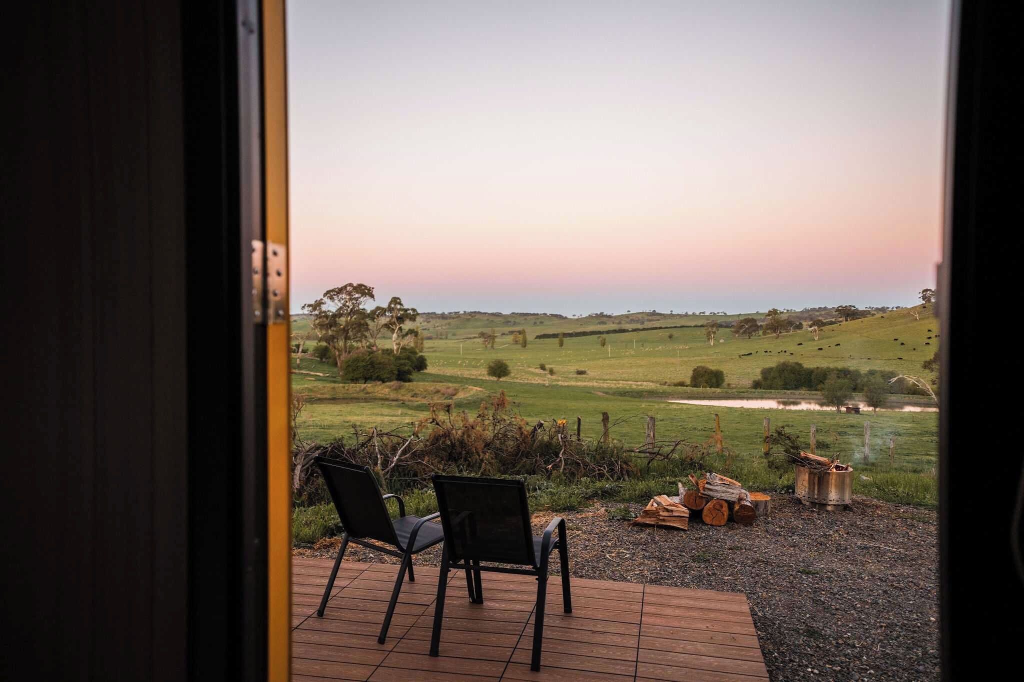 A patio with two chairs and a fire pit, overlooking a grassy field with trees and a hill in the distance.