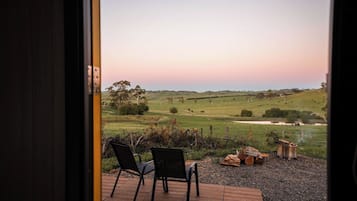 A patio with two chairs and a fire pit, overlooking a grassy field with trees and a hill in the distance.