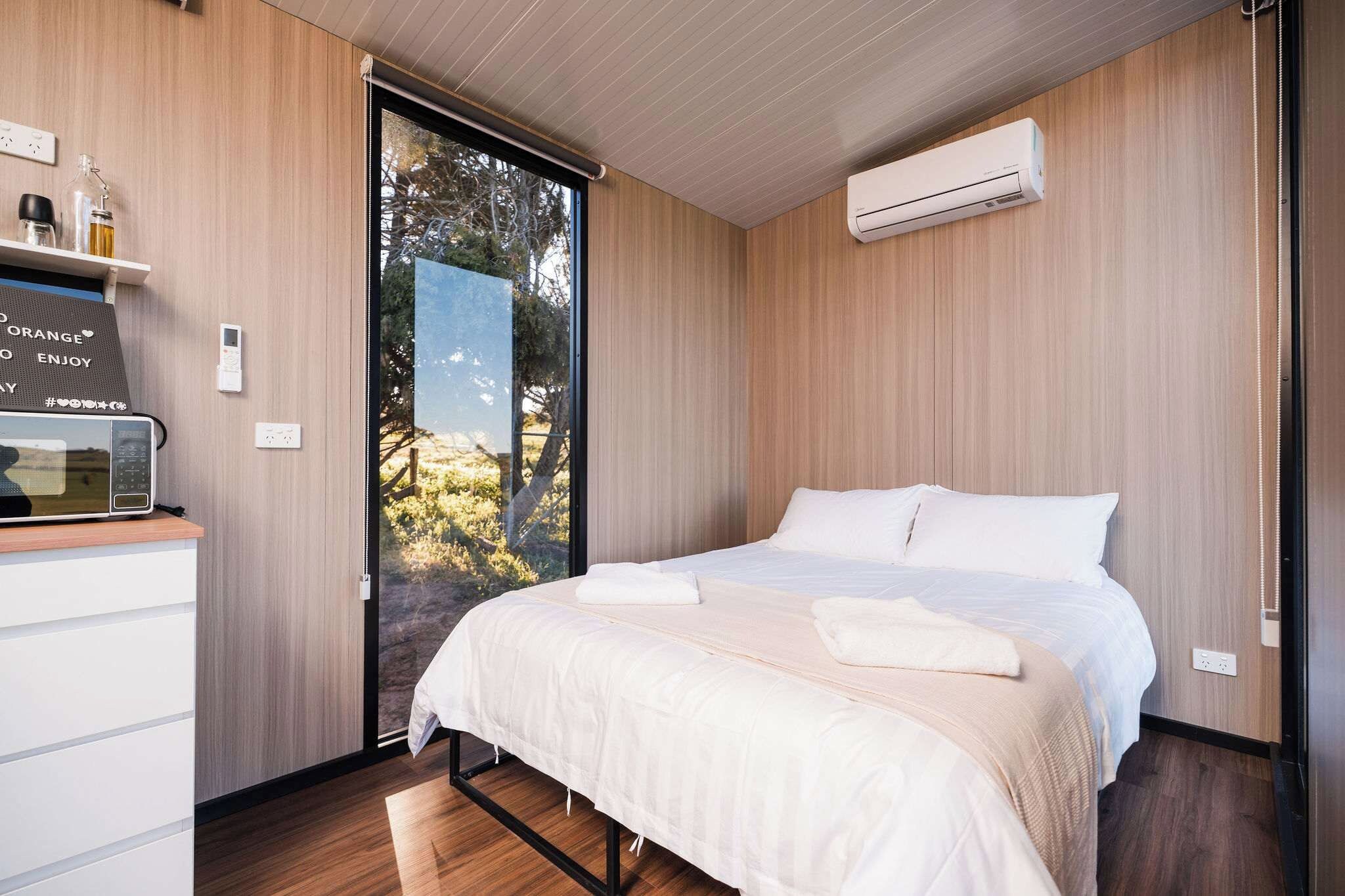 Un dormitorio moderno con cama, aire acondicionado integrado, una mesita con hervidor y vistas a través de un ventanal.