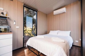 A modern bedroom with a bed, a built-in air conditioner, a small table with a kettle, and a view through a glass door.