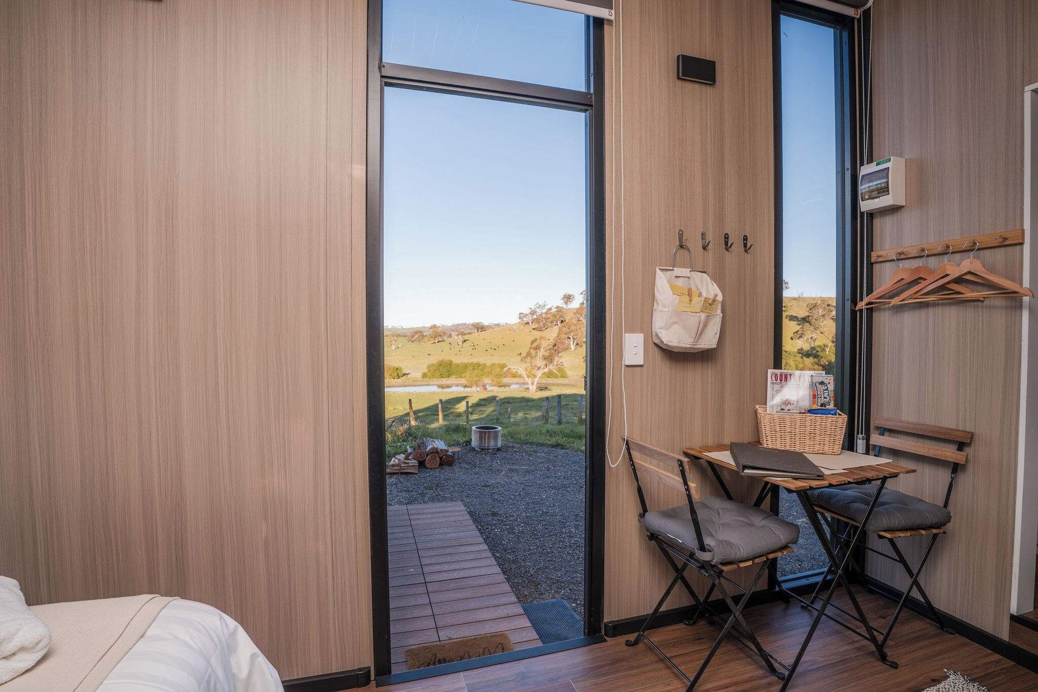 A room with a bed, a table with chairs, a view of a grassy landscape, and a wooden wall.