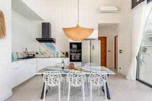 A modern kitchen with white cabinetry, a marble backsplash, a hanging light fixture, and a dining area with white chairs. - Villa Mona | Stylish 3BR Pool Villa (Bo Put)