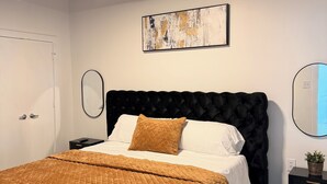 A modern bedroom with a black upholstered headboard, a quilted brown bedspread, a white pillow, a black nightstand with a plant, and a painting on the wall.