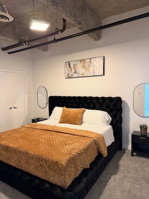 Un dormitorio moderno con un cabecero tapizado negro, una colcha marrón acolchada, una almohada blanca, una mesita de noche negra con una planta y un cuadro en la pared.