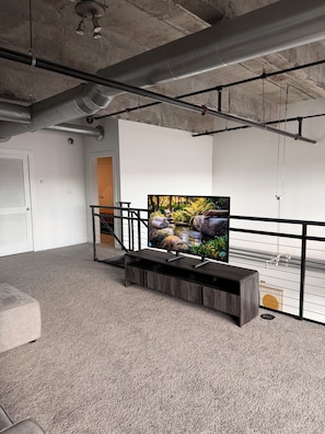 Amplia habitación con una gran televisión de pantalla plana, una escalera y un techo de concreto.
