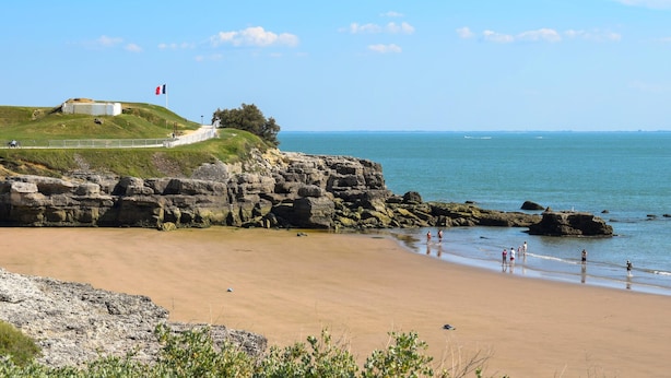 Een kustlandschap met een zandstrand, rotskliffen en een grasheuvel met een vlag.