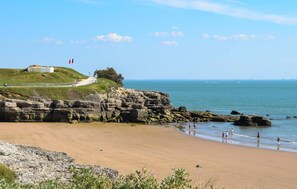 A coastal landscape with a sandy beach, rocky cliffs, and a grassy hill with a flag. - Charmante maison proche plage (La Tremblade)