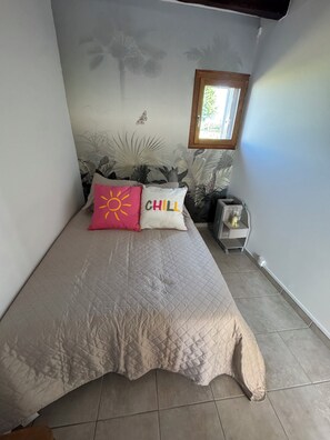 A bed with a quilted cover, two pillows with text, a small table with a glass, and a window with a view of greenery. - Vrbo Property (Valençay)