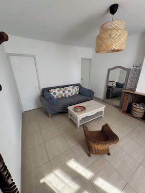 Een woonkamer met een bank, een bijzettafeltje, een rieten fauteuil, een hanglamp en een open haard.