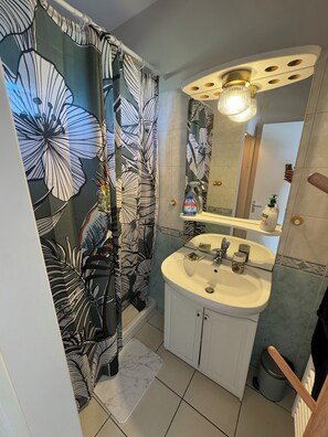 A bathroom with a floral shower curtain, a white sink with a mirror above it, and a handheld showerhead. - Vrbo Property (Valençay)