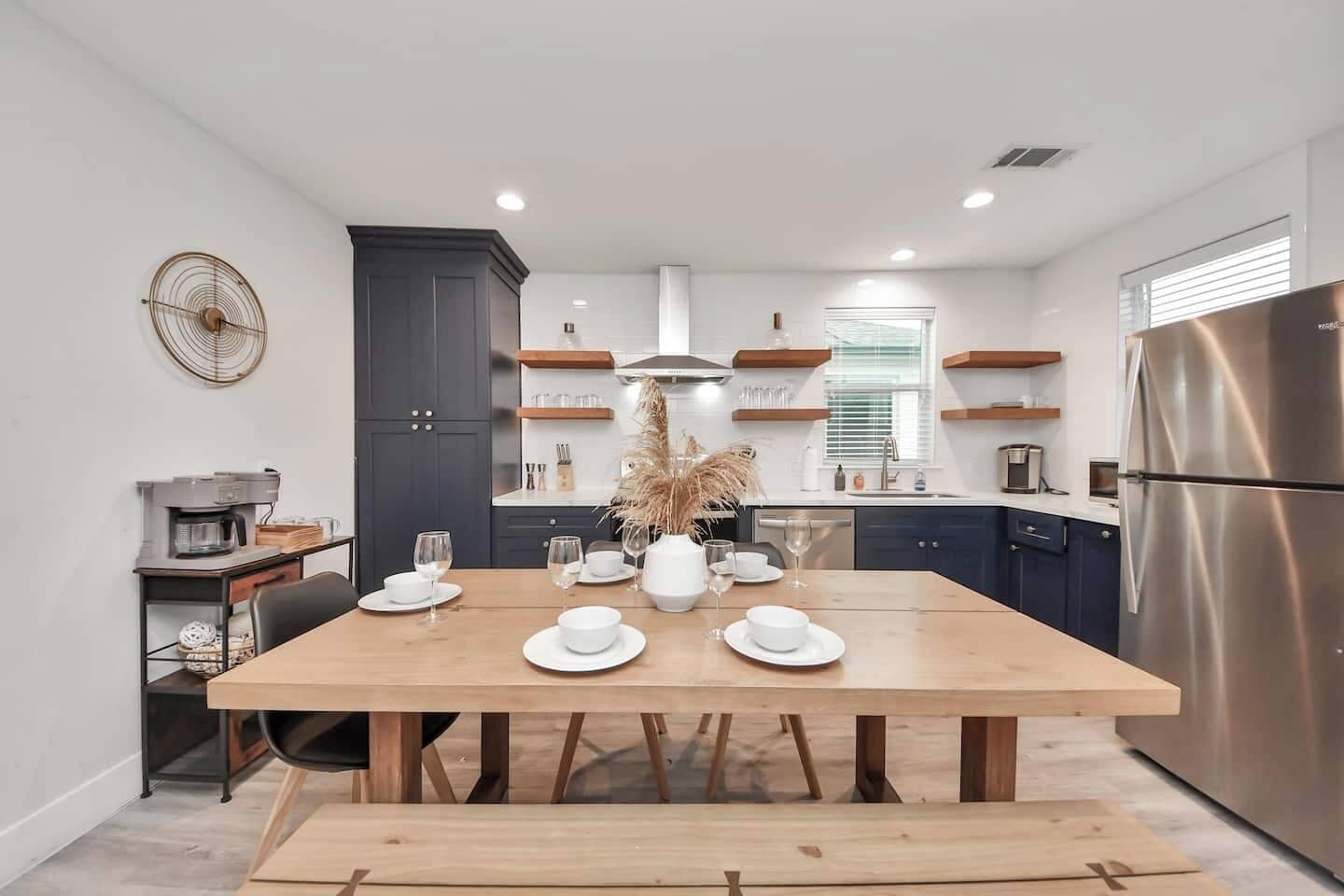Une cuisine moderne avec des meubles de couleur sombre, une table à manger en bois agrémentée d’assiettes et de verres blancs, un réfrigérateur en acier inoxydable et une machine à café.