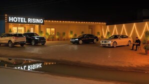 Ein Hotel namens 'HOTEL RISING' mit einer gut beleuchteten Fassade, einem Parkplatz und einer Person, die draußen auf einem Stuhl sitzt.