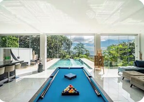 Et billardbord i et moderne rum med store vinduer, et barområde og udsigt til en pool og grøn bevoksning.