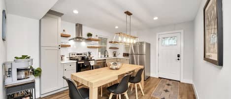 A modern kitchen with a wooden dining table, black chairs, a stainless steel refrigerator, and a hanging light fixture.