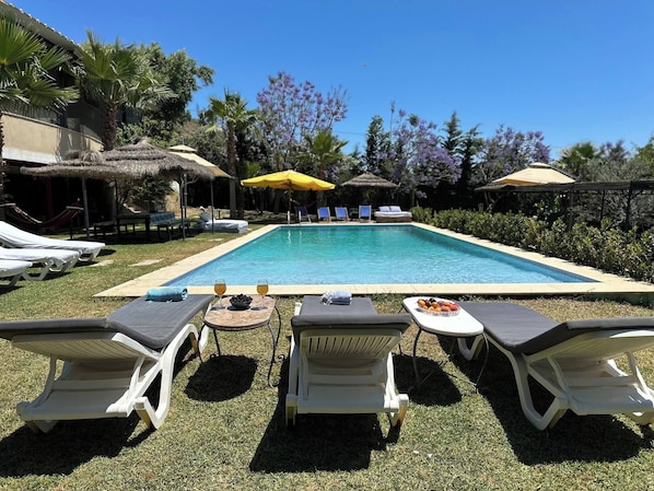 A swimming pool with lounge chairs and umbrellas, surrounded by greenery and a clear sky. - Grotte de Sel Private Pool (Ghar El Melh)