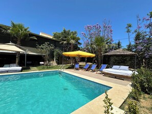 Une piscine entourée de verdure et d’un bâtiment, avec des chaises longues et des parasols.