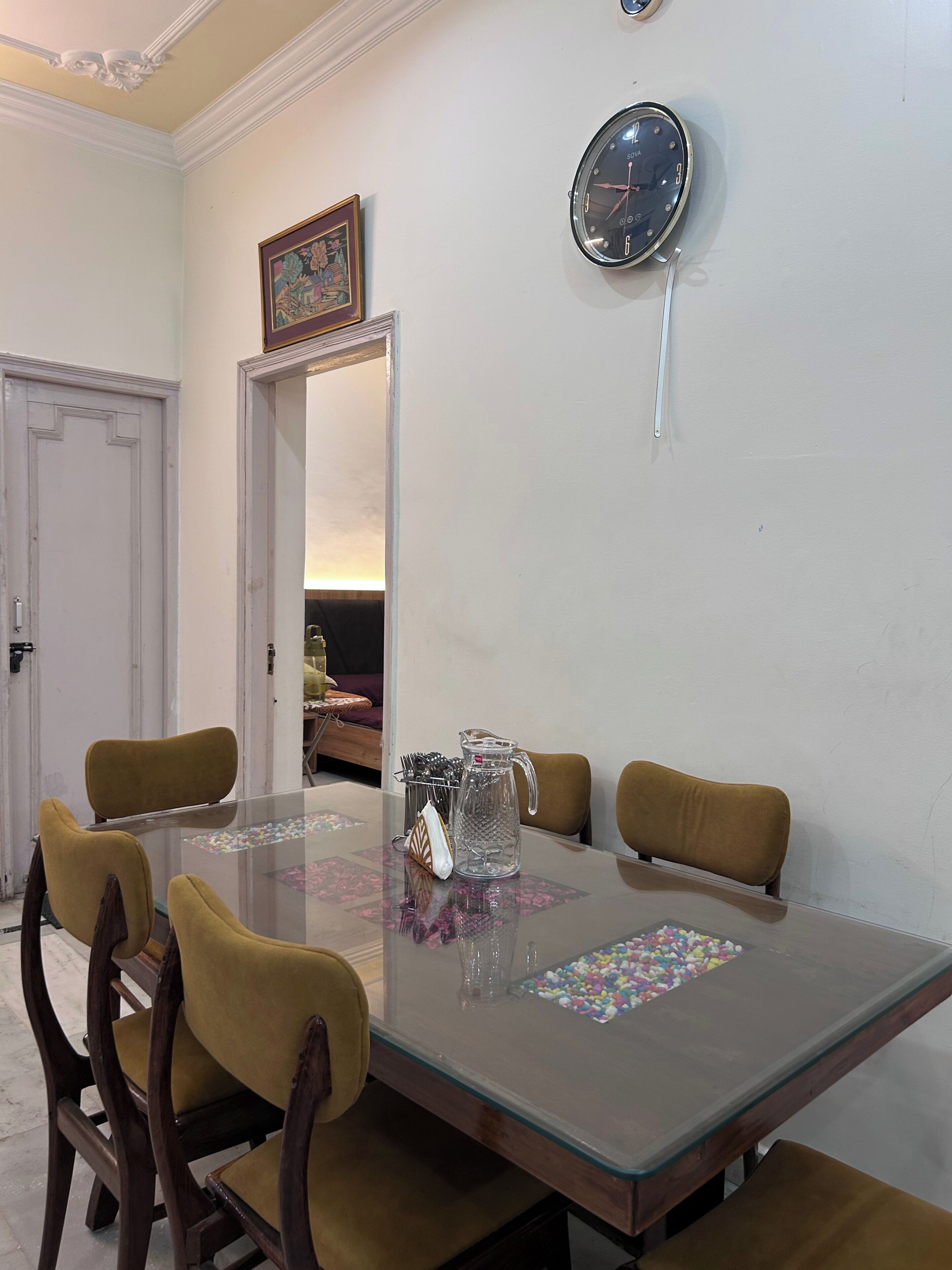 A dining area with a glass table, wooden chairs, a pitcher, and a clock on the wall.