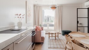 A modern kitchen with a countertop, a sofa, a dining table with chairs, and a view of buildings through large windows. - Premium 3BR Home | Prime Location | Modern Touch (Helsinki)