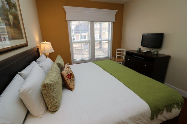 A hotel room with a bed, a television, a window with blinds, and a painting on the wall.