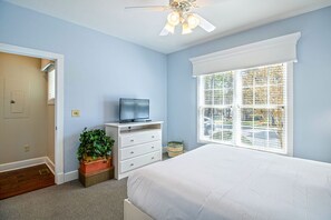 Un dormitorio con cama, televisor sobre una cómoda, ventana con persianas y ventilador de techo.