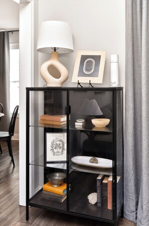 Um armário de vidro preto com prateleiras que contêm livros, uma tigela, um quadro emoldurado e uma luminária.
