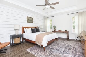 Quarto com cama, mesas de cabeceira, ventilador de teto, janela com cortinas e vista para o verde lá fora.