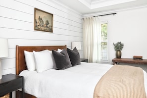 Un dormitorio con cabecera de madera, ropa de cama blanca, una mesita de noche con lámpara, un cuadro enmarcado en la pared y una ventana con cortinas.