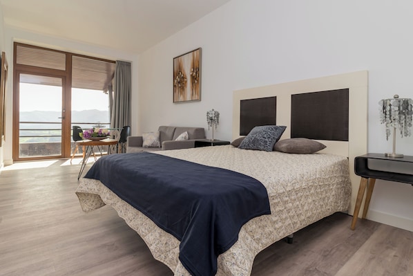 A bedroom with a large bed, a desk, a chair, a balcony, and a view of the mountains.