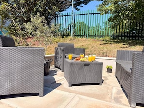 Outdoor wicker furniture with a table set for two, surrounded by a paved patio area. - Unique Devon Cottages - Sunrise Cottage - Stylish & Comfortable (Exmouth)