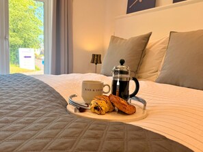 A bed with a quilted bedspread, a tray with a coffee pot, a mug, and pastries. - Unique Devon Cottages - Sunrise Cottage - Stylish & Comfortable (Exmouth)